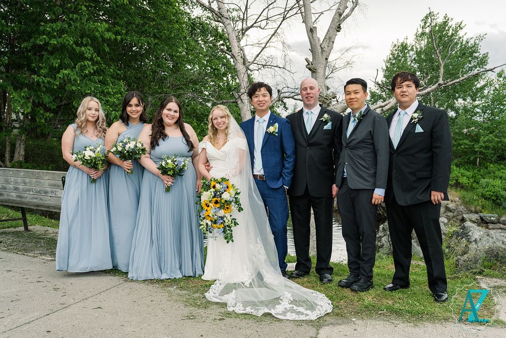 Full wedding party in blue and black formal attire poses outdoors with bridesmaids in light blue dresses.