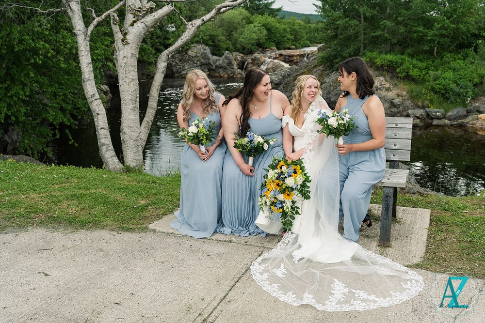 Bridal party in light blue dresses poses with bride holding sunflower bouquet by water.