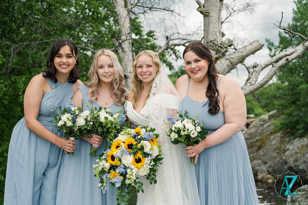 Wedding party in light blue bridesmaid dresses holding white and yellow floral bouquets against a natural outdoor backdrop.