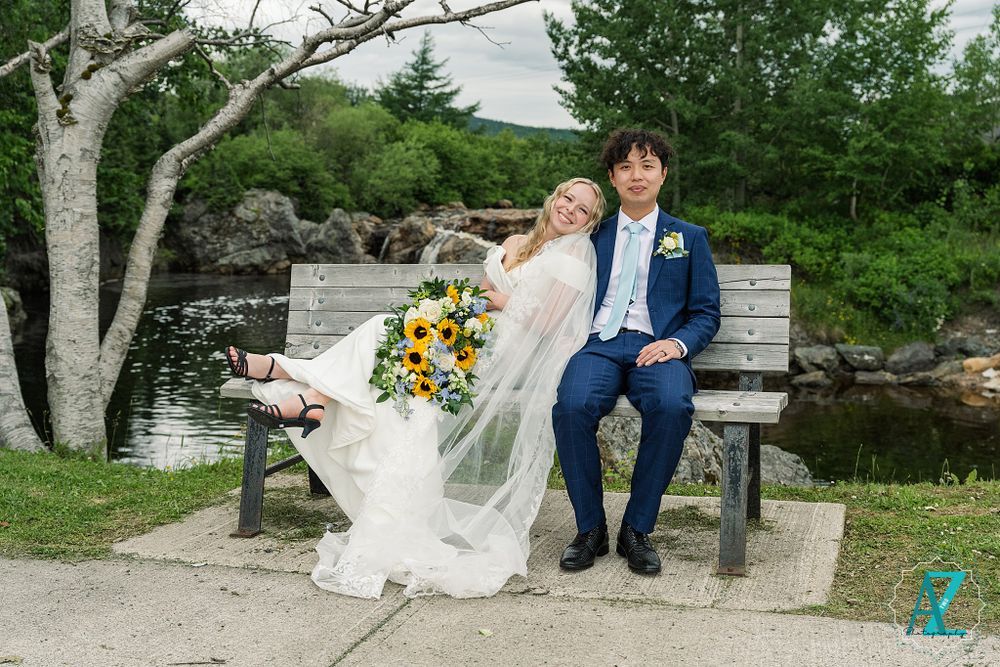 Newlyweds sitting on a park bench by a pond with flowing white veil and sunflower bouquet.