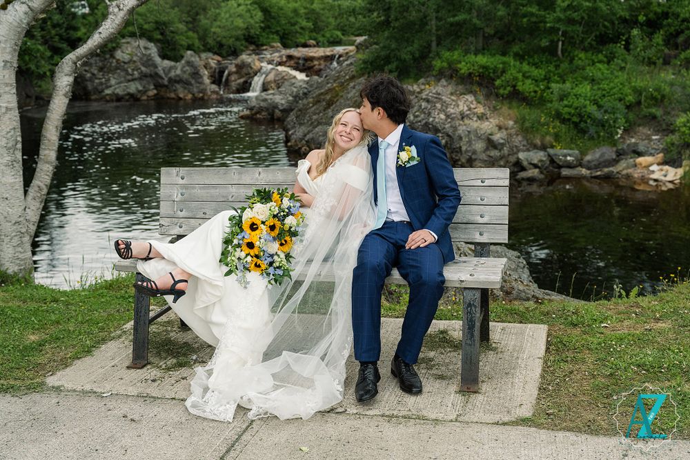 Couple sitting on stone bench near water sharing a romantic moment with flowing veil and sunflower bouquet.