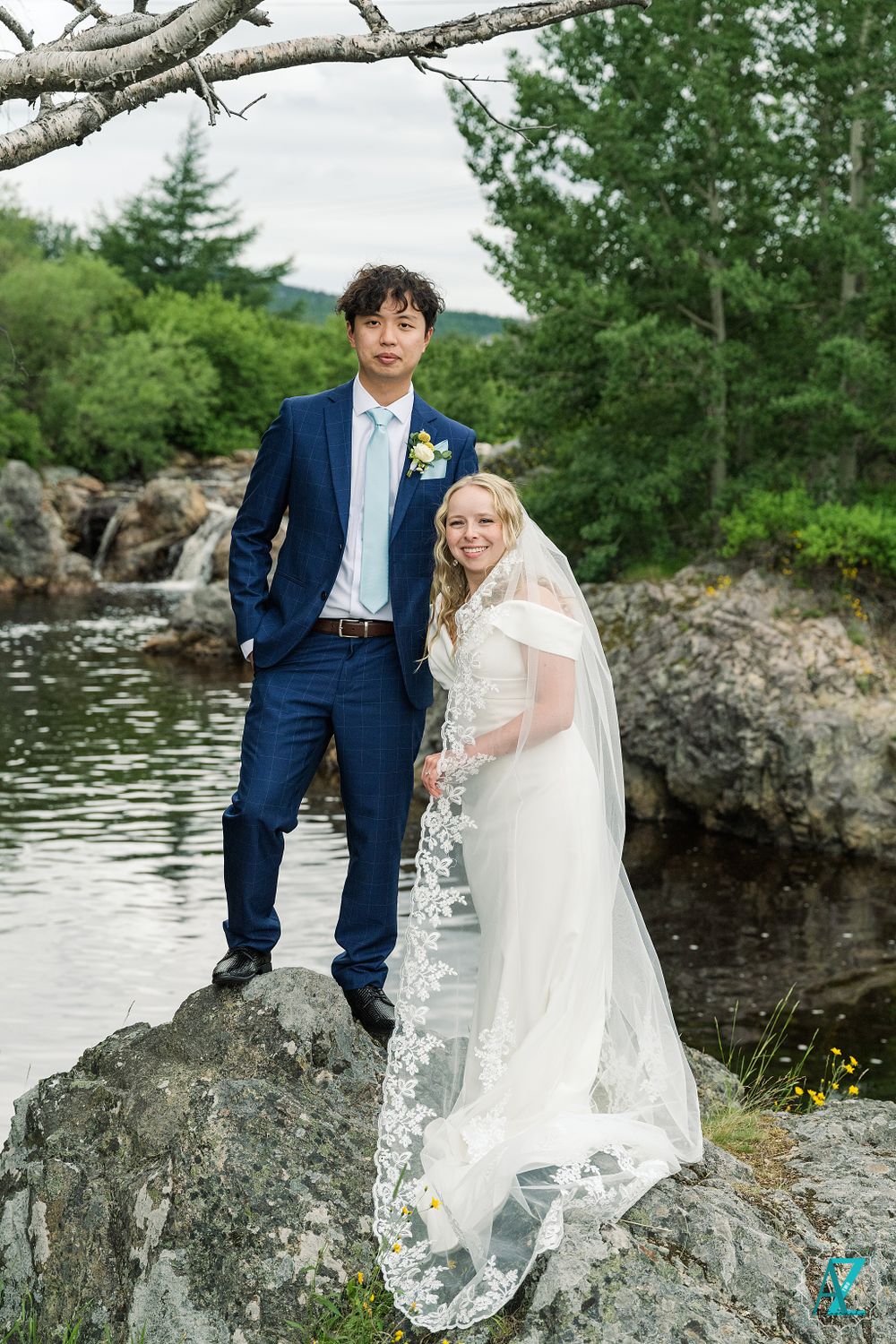 Wedding couple posing on rocky outcrop by water with long flowing veil and navy suit.
