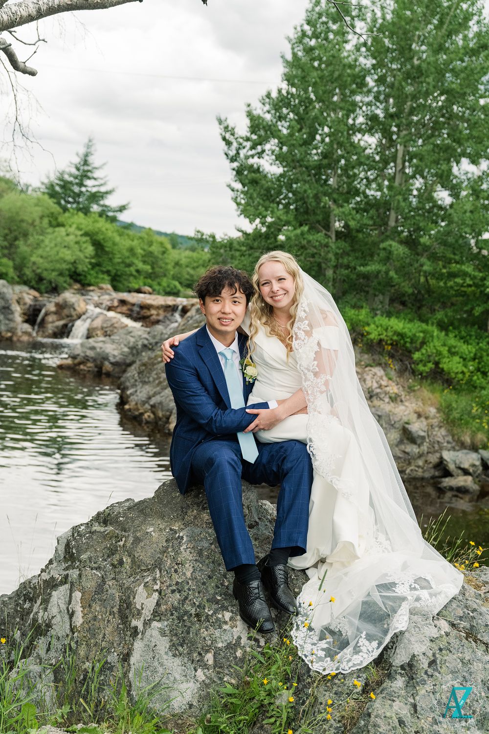 Couple sitting on rocky ledge overlooking water in elegant wedding attire.