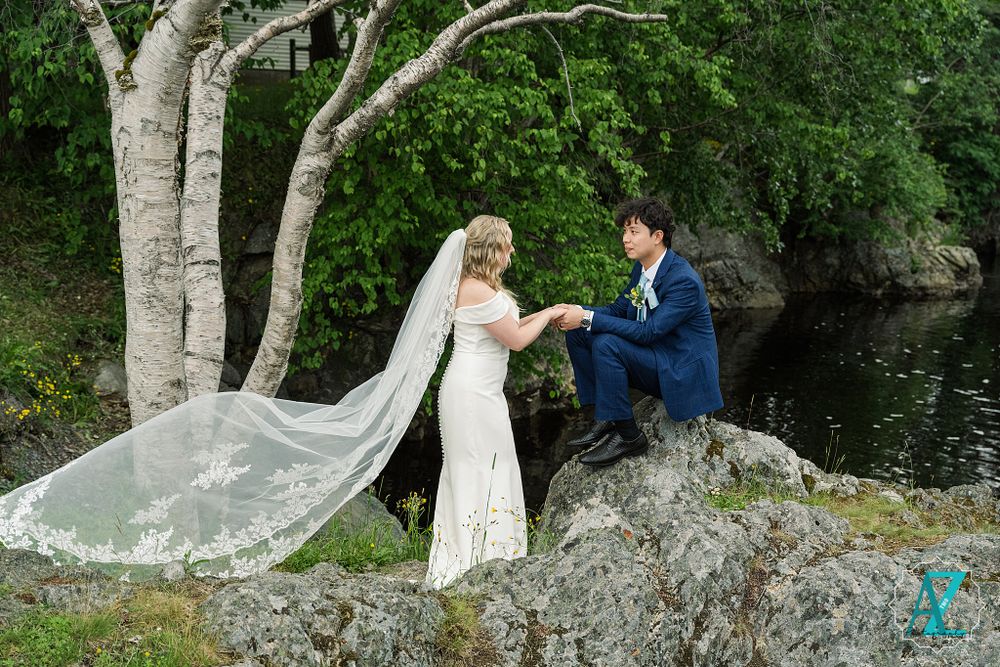 Wedding couple on rocky outcrop with flowing veil against natural water backdrop.