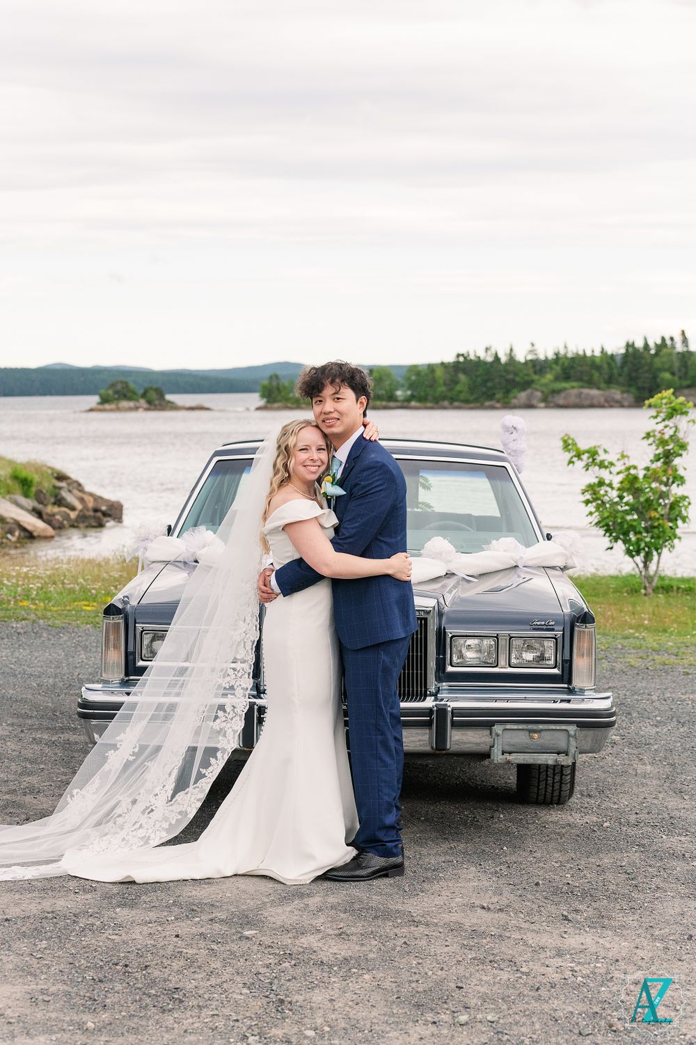 Couple in wedding attire pose in front of vintage car with scenic water view background.