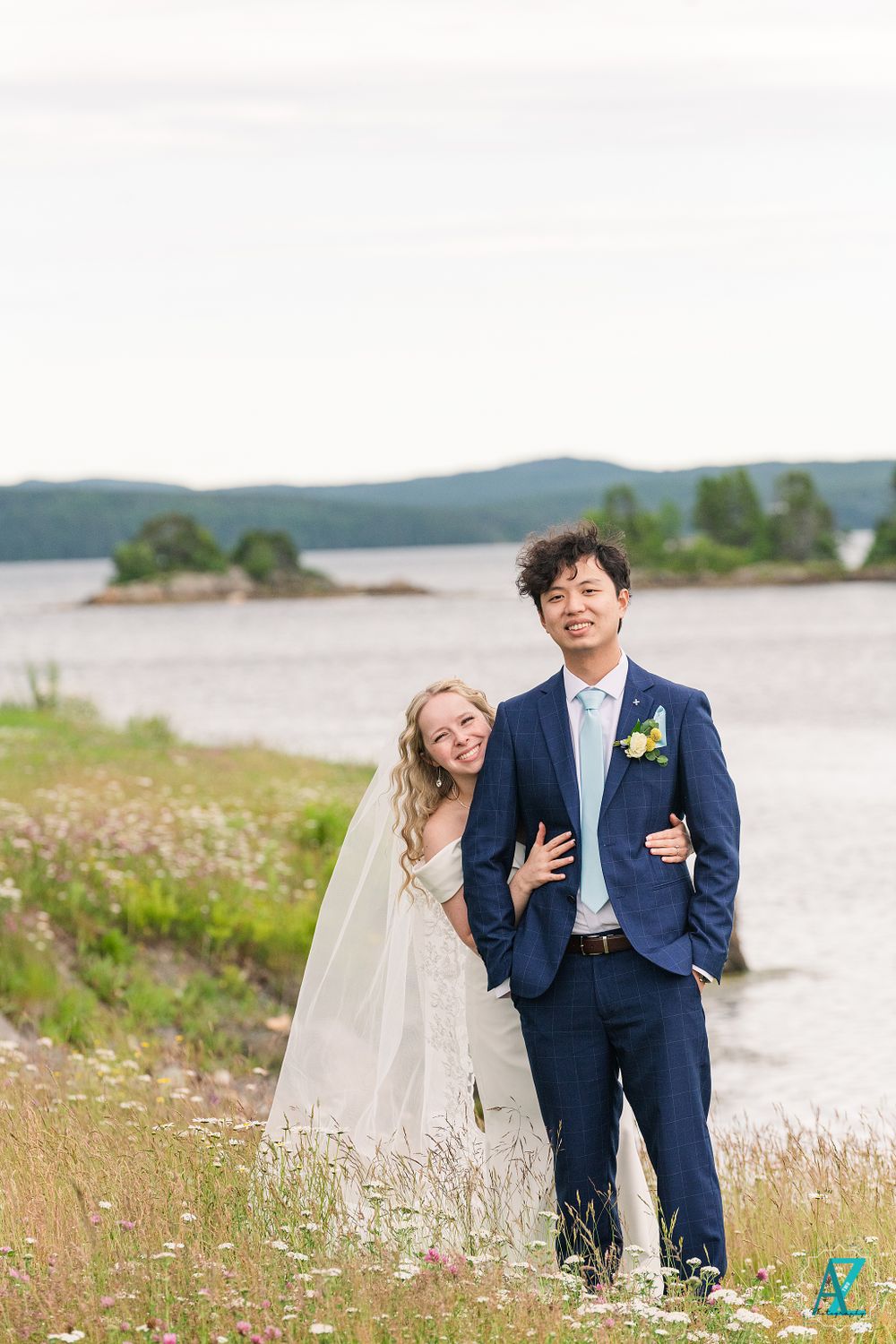 Newlyweds stand together on a grassy hillside overlooking a serene lake and mountains.