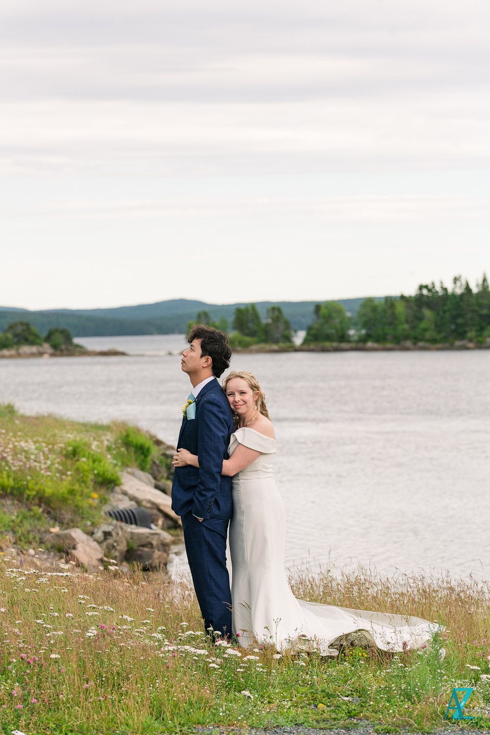Wedding couple poses back-to-back on a scenic lakeside with mountains in the background.
