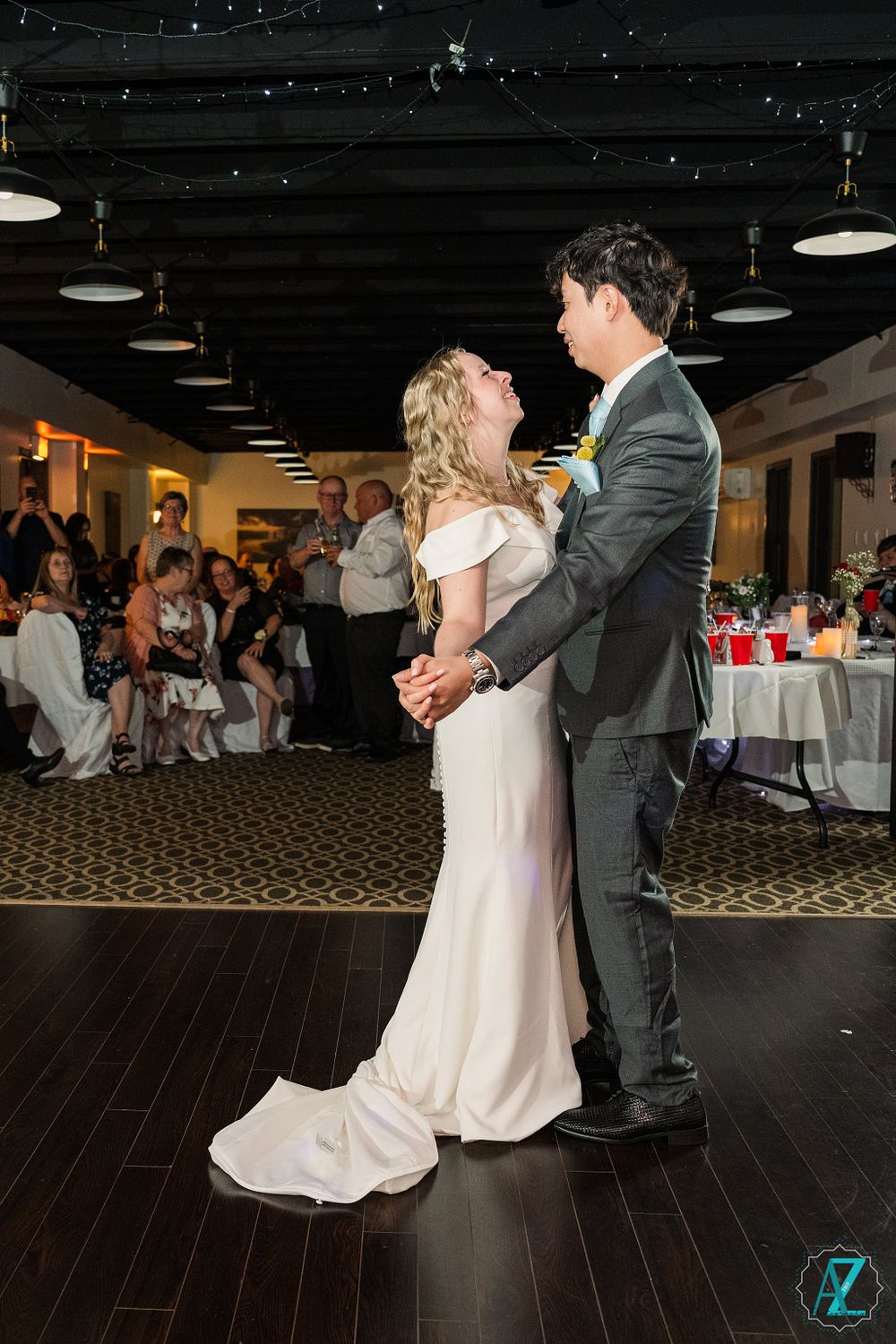 A couple shares their first dance at a wedding reception with guests and tables visible in the background.