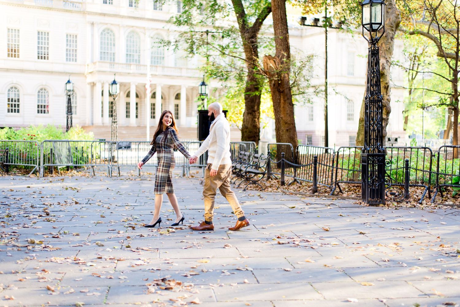 woman in plaid dress leads man in white sweater by hand at NYC City Hall in the fall