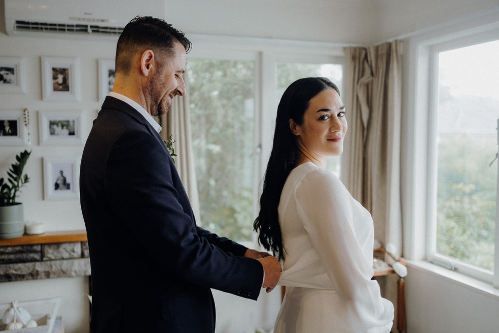 Intimate wedding preparation moment in bright room with large windows and photo wall backdrop.