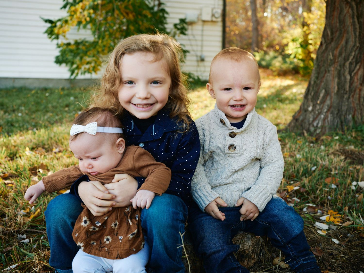 Three children sitting together and laughing among fallen autumn leaves.