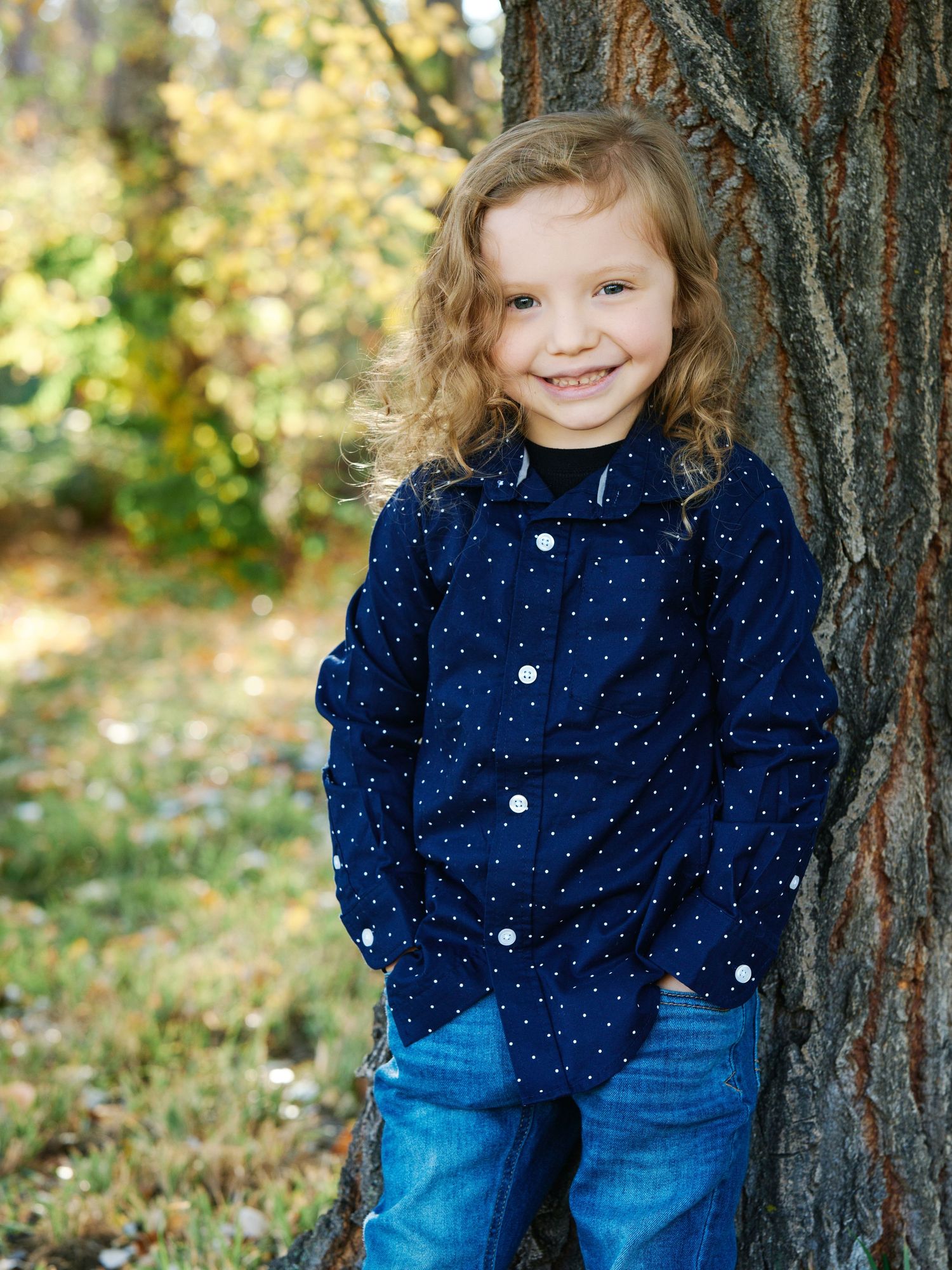 A child in a polka dot navy shirt leans against a tree trunk in an autumn setting.