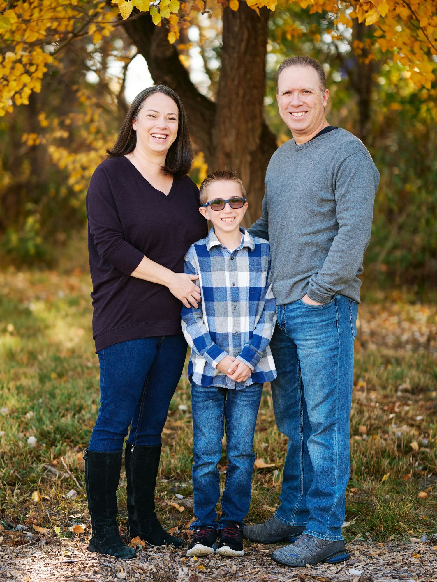 A family of three poses together against a backdrop of autumn trees.