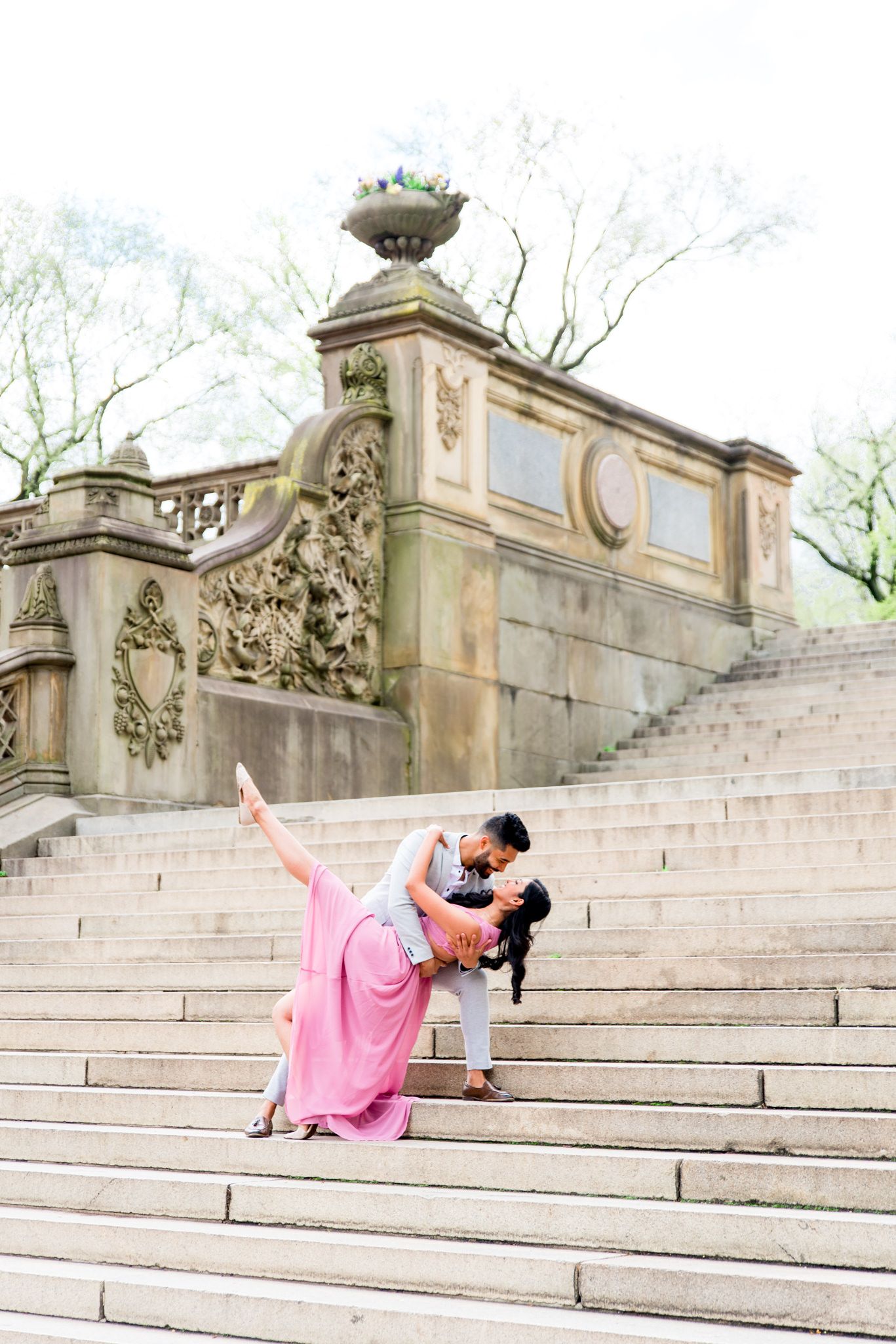 Woman in pink dress and man in gray suit dancing on steps to Bethesda Terrace Central Park for spring engagement photos