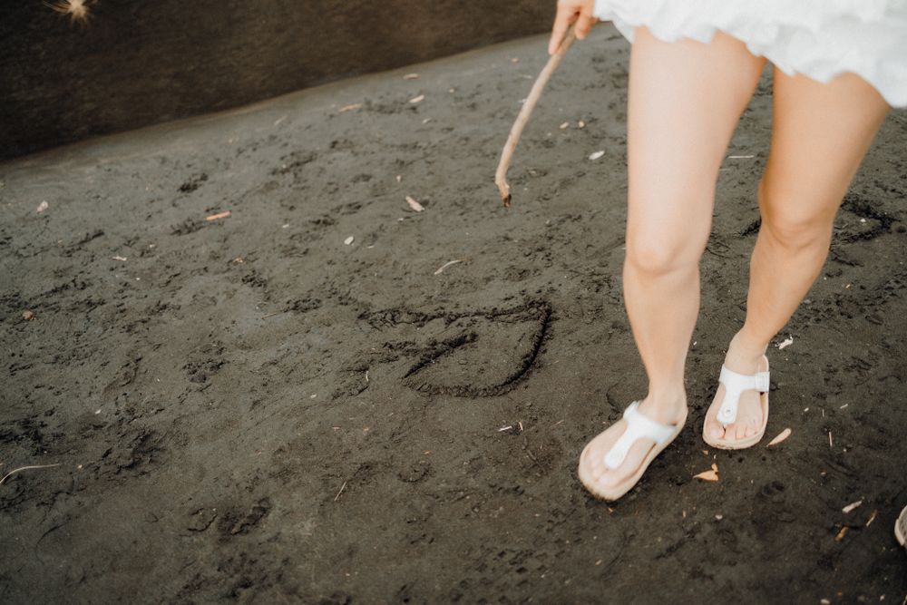 White shoes on black sand beach with someone drawing a heart shape with a stick.