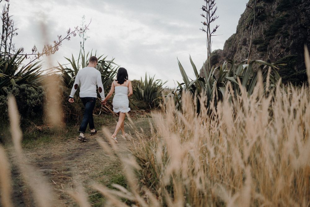 Two people walking on a coastal path with tall grass and mountains in the background.