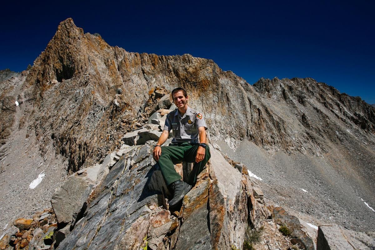 Jeff the park ranger taking a break on a rocky mountain ridge against a deep blue sky and rugged peaks.