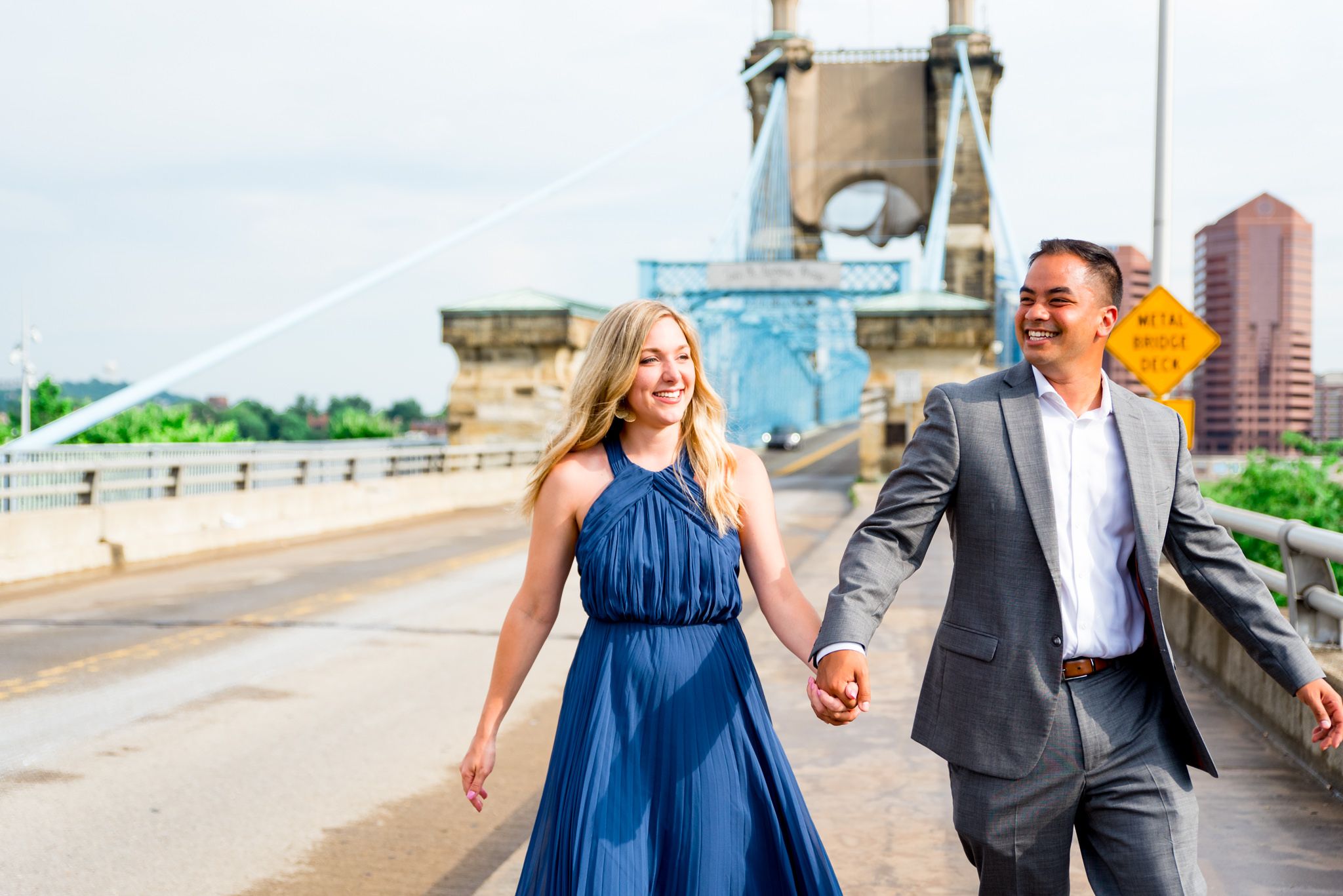 man in grey suit holds hand of blonde woman in blue dress as they walk near the Roebling Bridge for engagement pictures