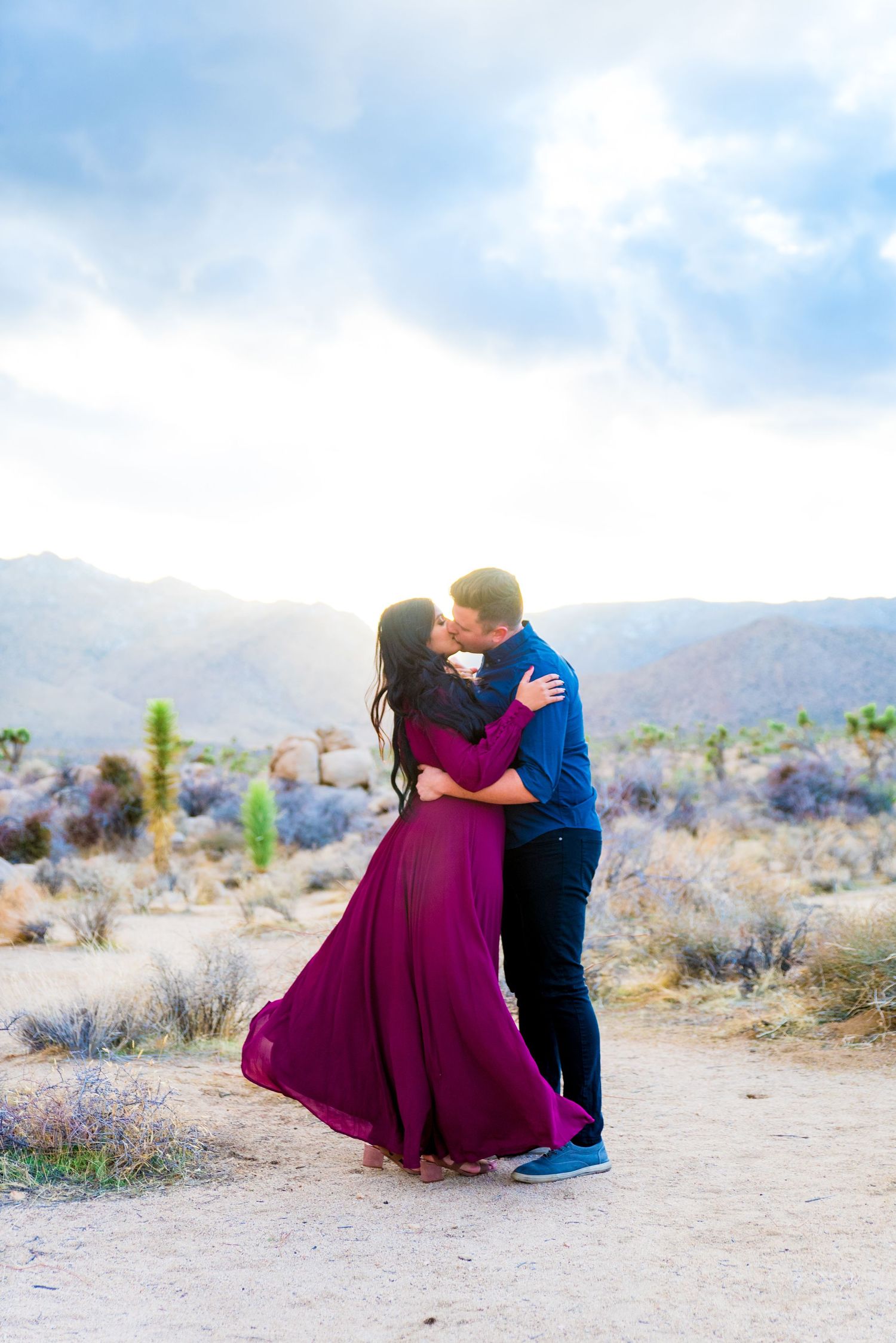 woman with long dark hair in red dress kissing man in blue shirt under the sunset at Joshua Tree National Park