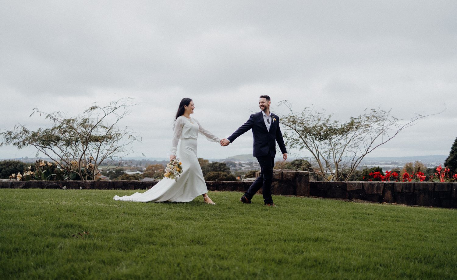 Couple holding hands while walking on grass with cloudy sky behind them.