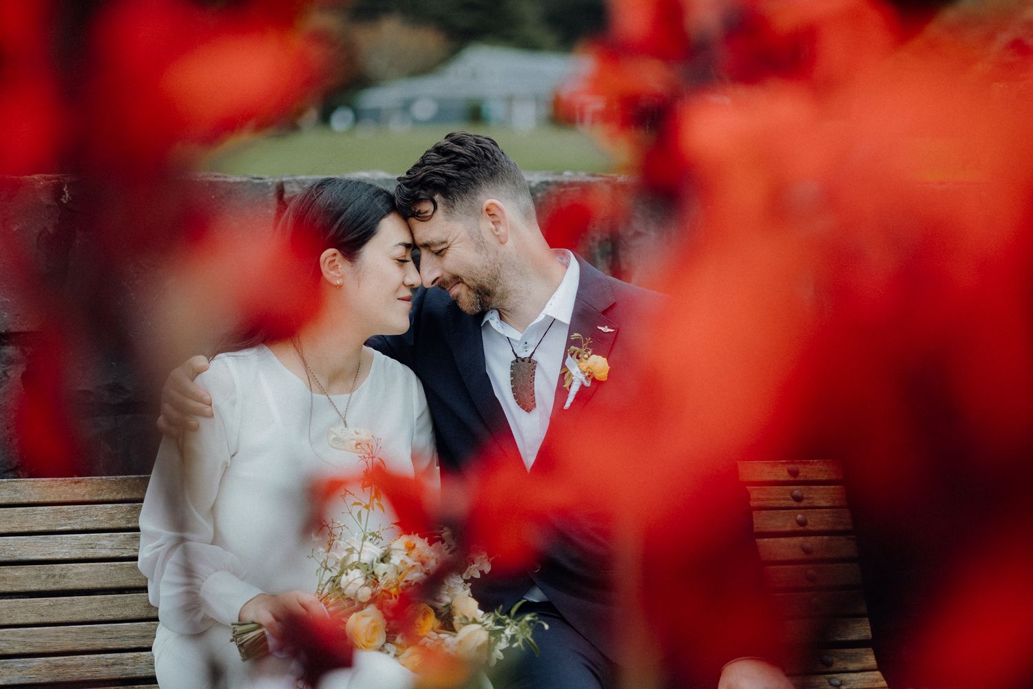 A romantic moment captured through red flowers with a couple sitting on a wooden bench in an outdoor setting.