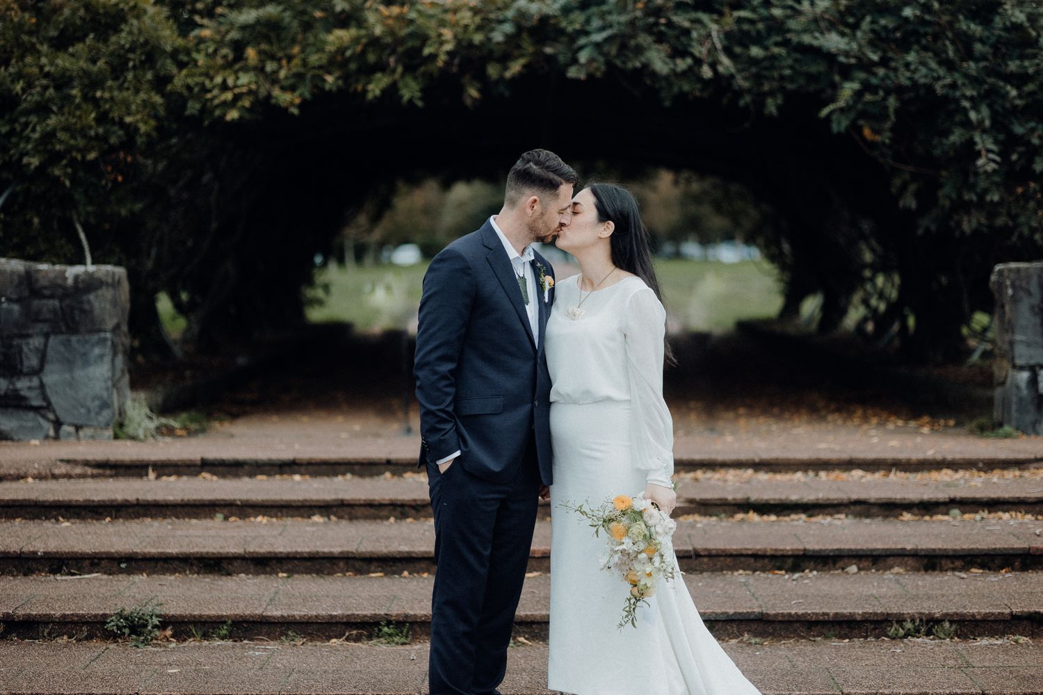 A couple poses beneath a garden archway on stone steps in elegant wedding attire.