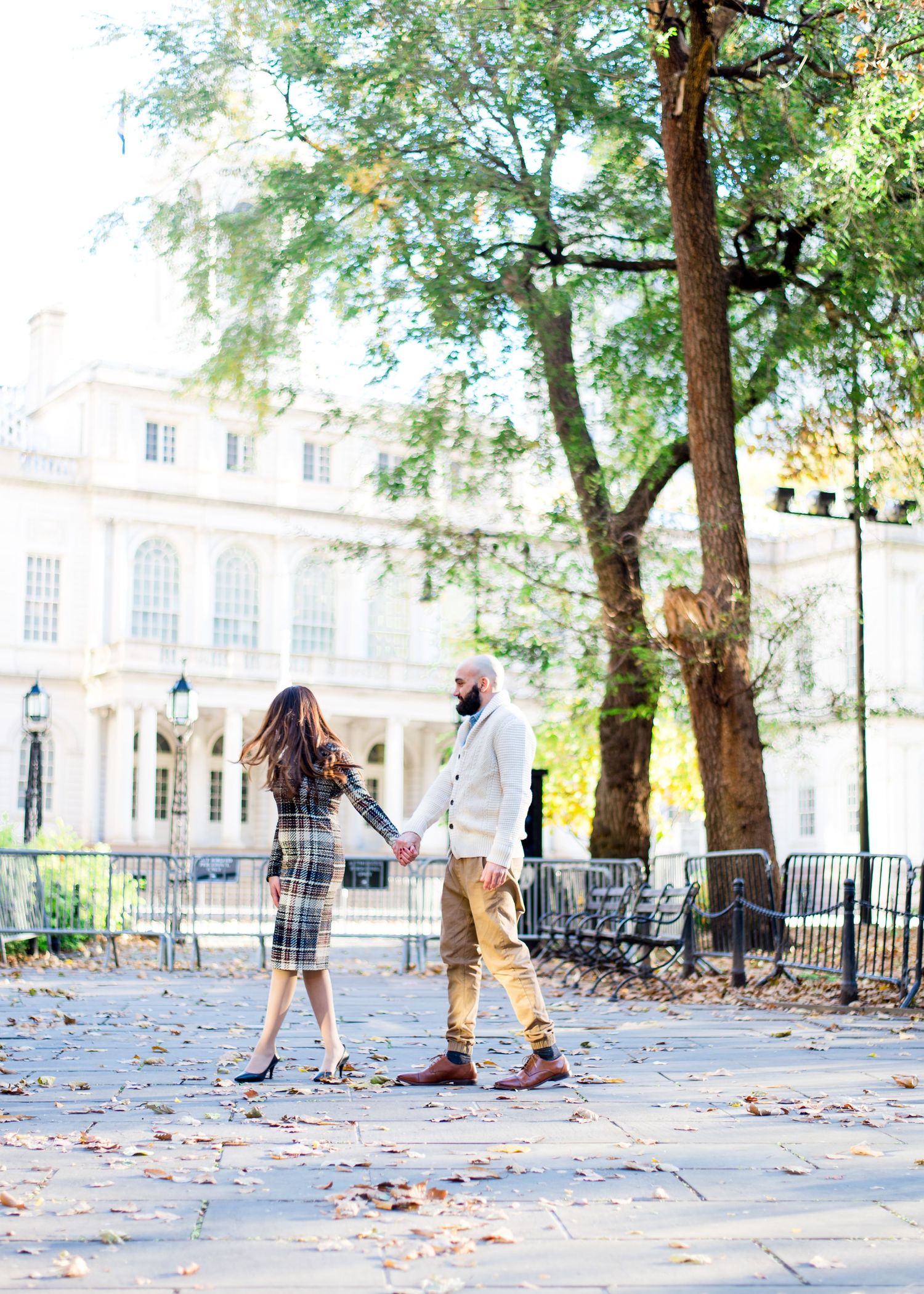 woman in plaid dress holds hand of man in white sweater in front of New York City Hall for engagement pictures