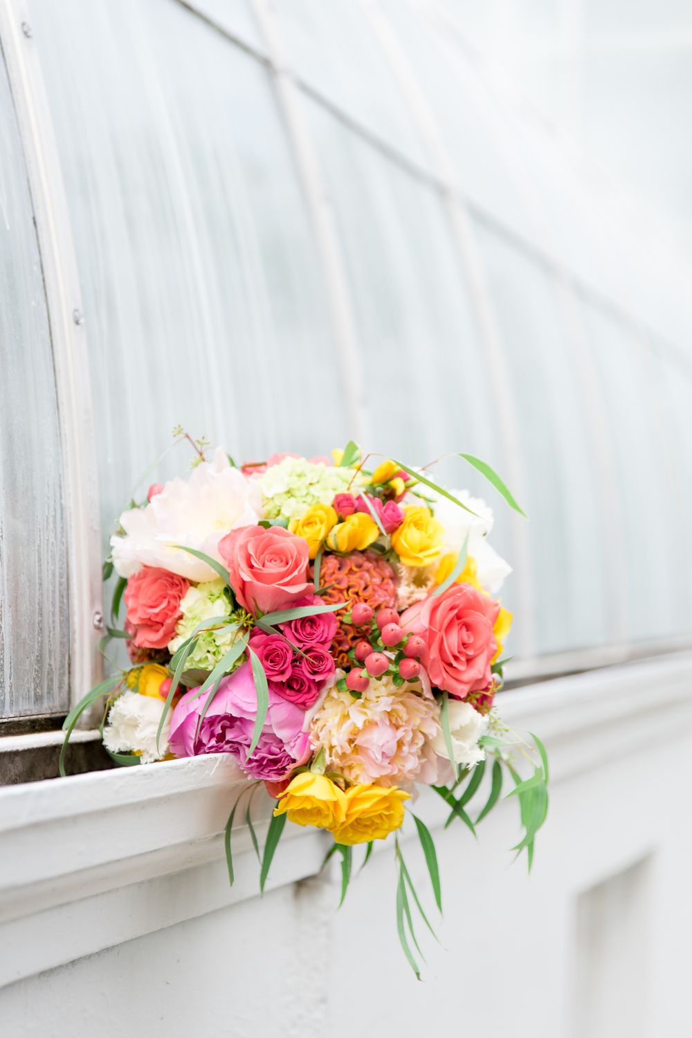 summer wedding bouquet with coral, pink, and yellow mixes roses and peonies against a glass conservatory window