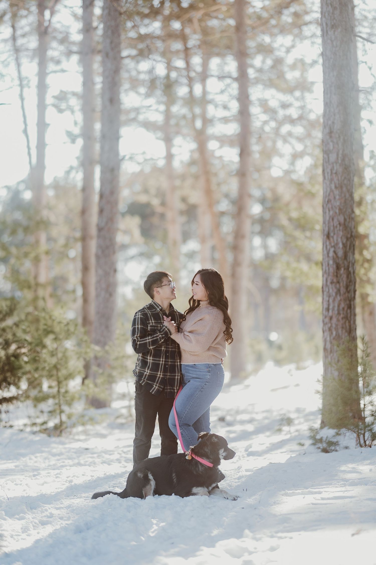 Couple shares intimate moment in snowy forest with their dog sitting below.