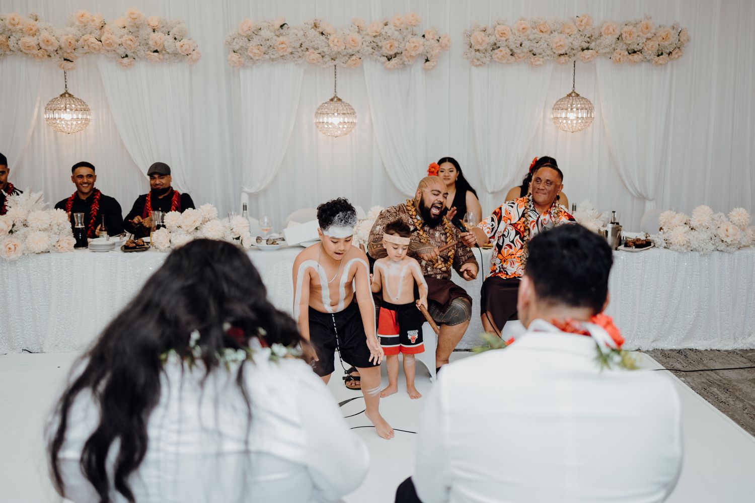 Wedding celebration features energetic dancing in front of elegant white floral wall decor.