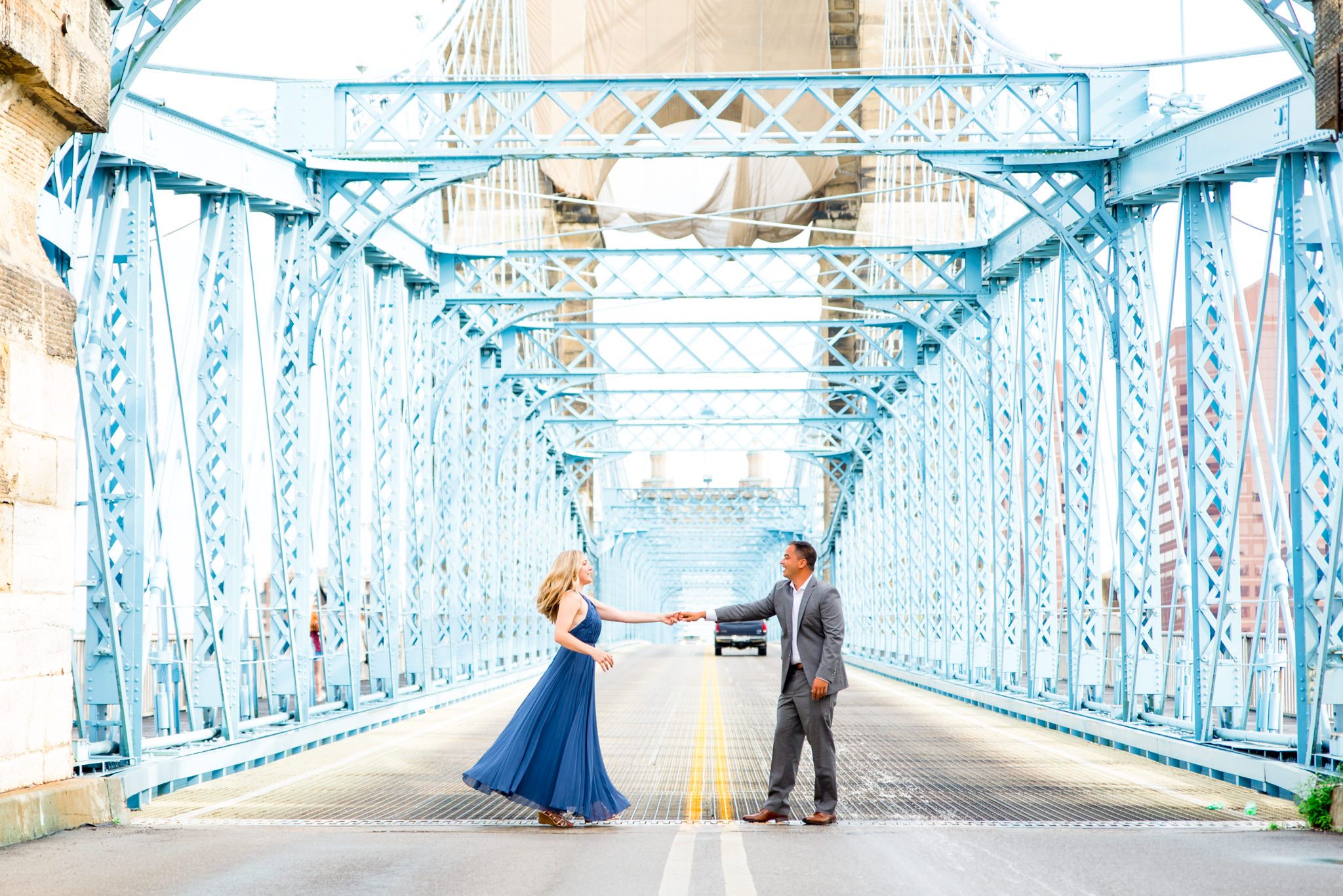 man in gray suit holds hand of woman in long blue dress on Roebling Bridge engagement pictures