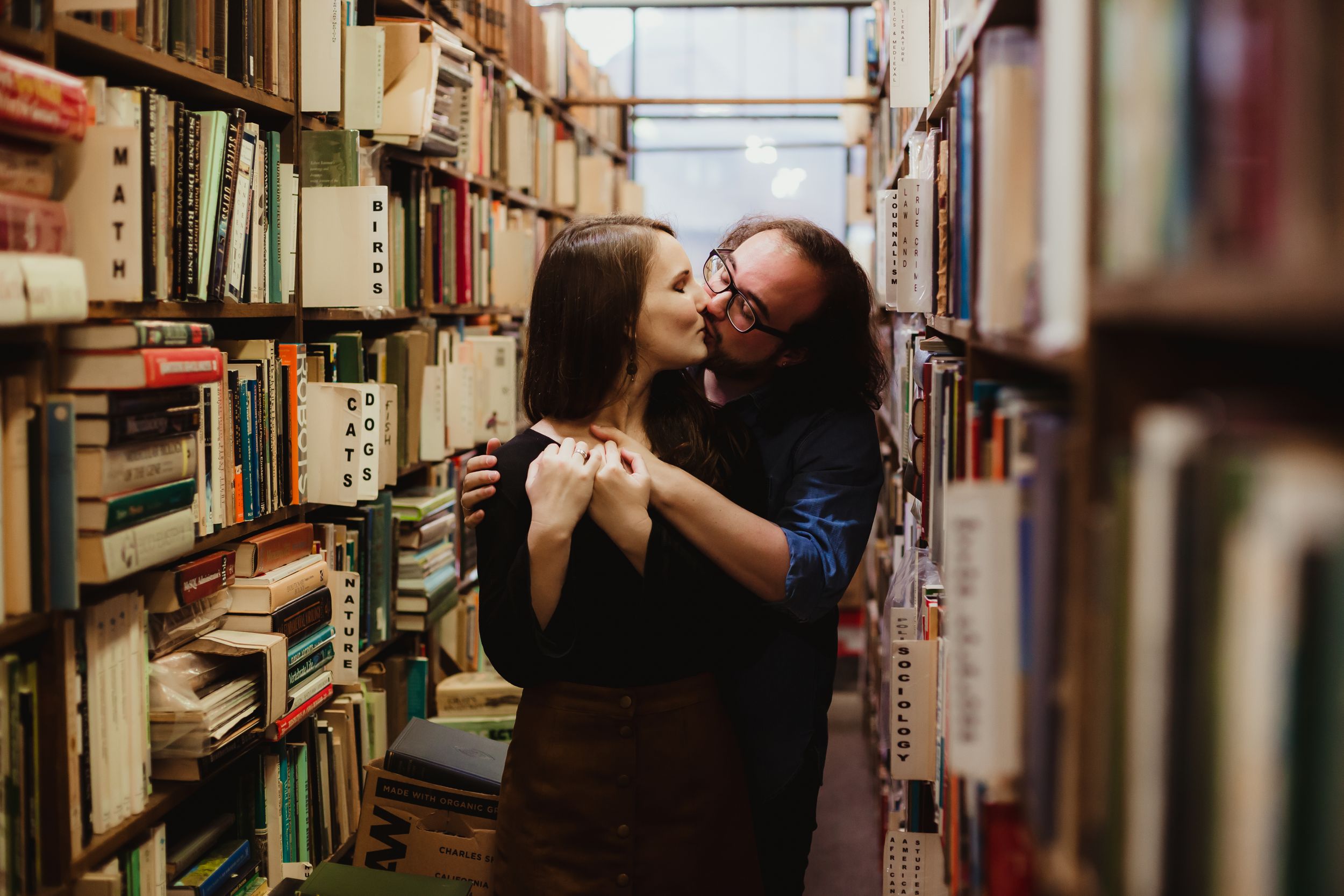 An engaged man and woman with brown hair kissing each other in between shelves of old books at a store.