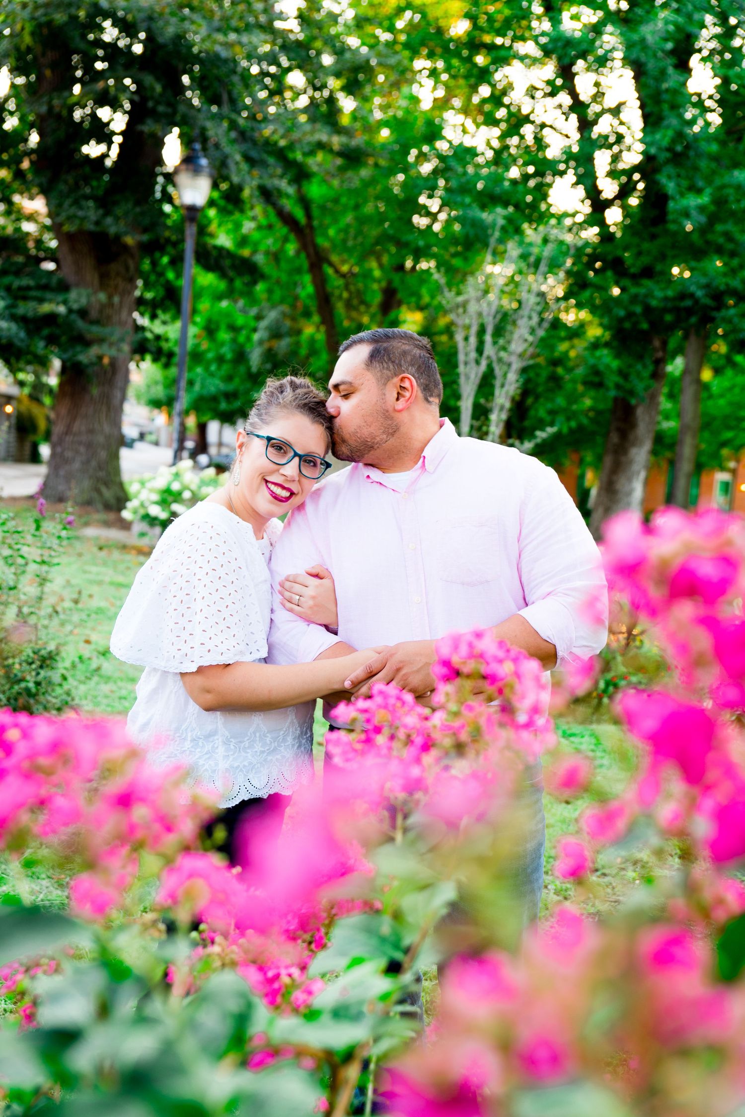 man kisses his fiancee's forehead as she holds him and smiles standing behind a pink flower bush