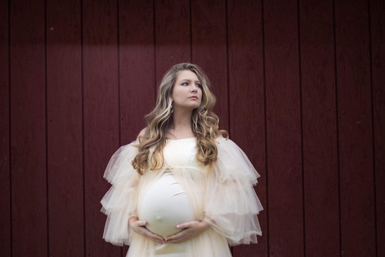 Ethereal maternity portrait featuring a cream-colored tulle dress against a rustic red barn wall.