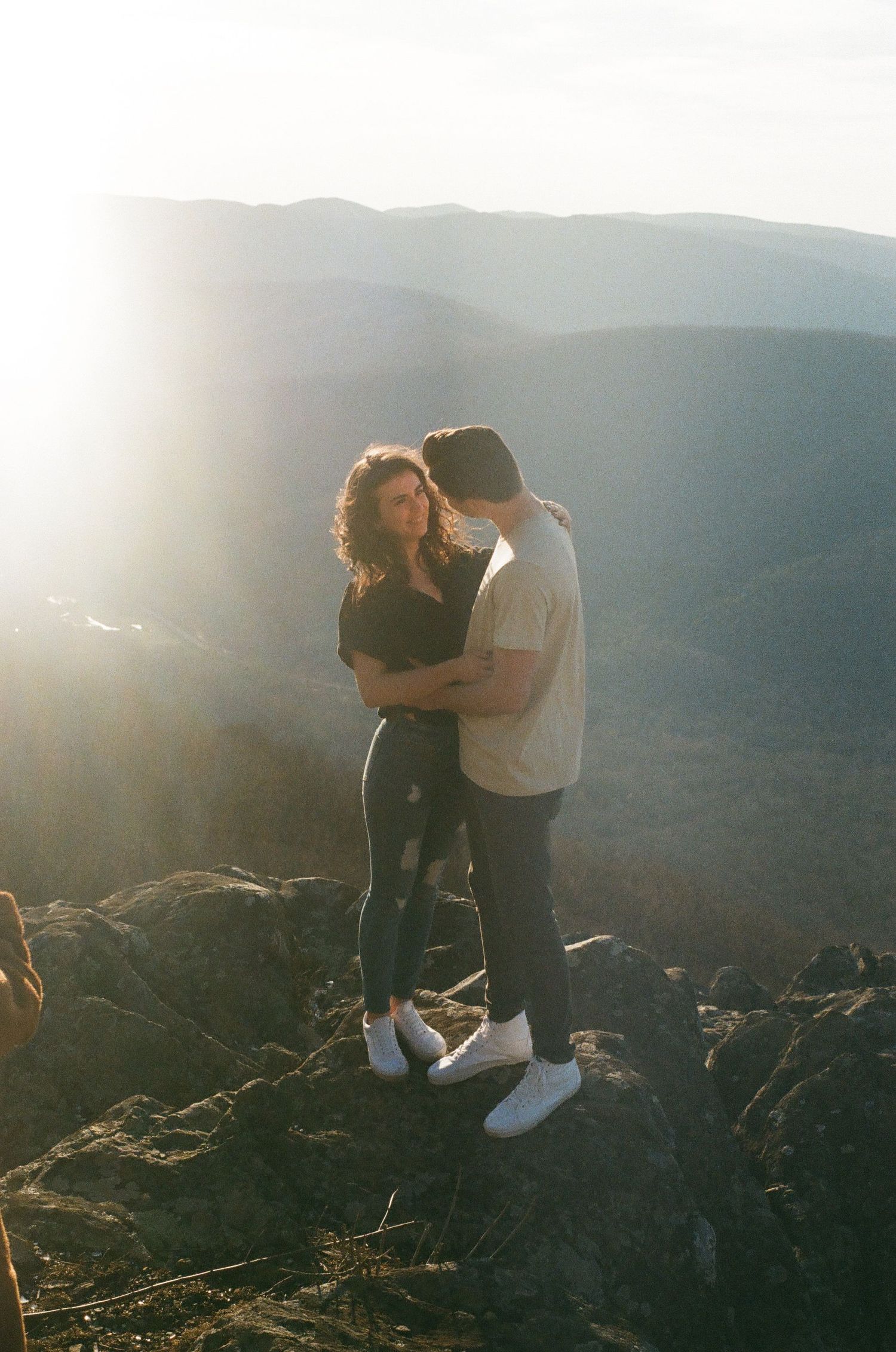 Couple sharing an intimate moment at sunset on a mountaintop overlooking a scenic mountain range.