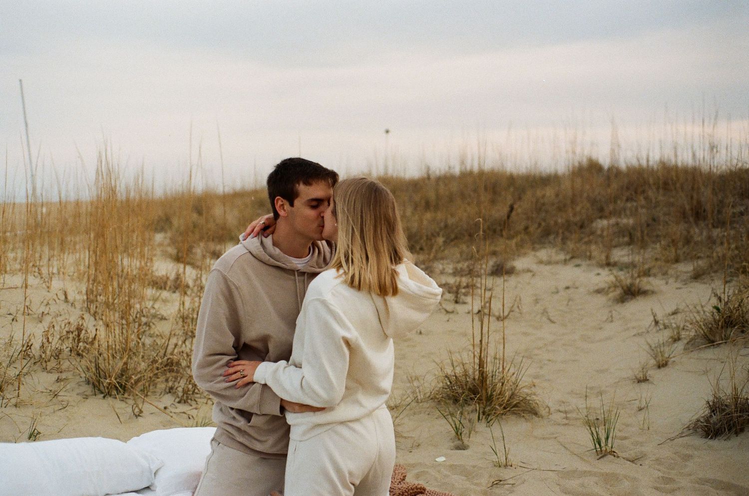 A couple embraces on sand dunes during golden hour with beach grass in the background.