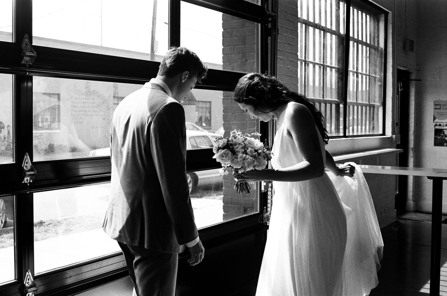 Black and white photo of couple by window with wedding attire examining floral arrangements.