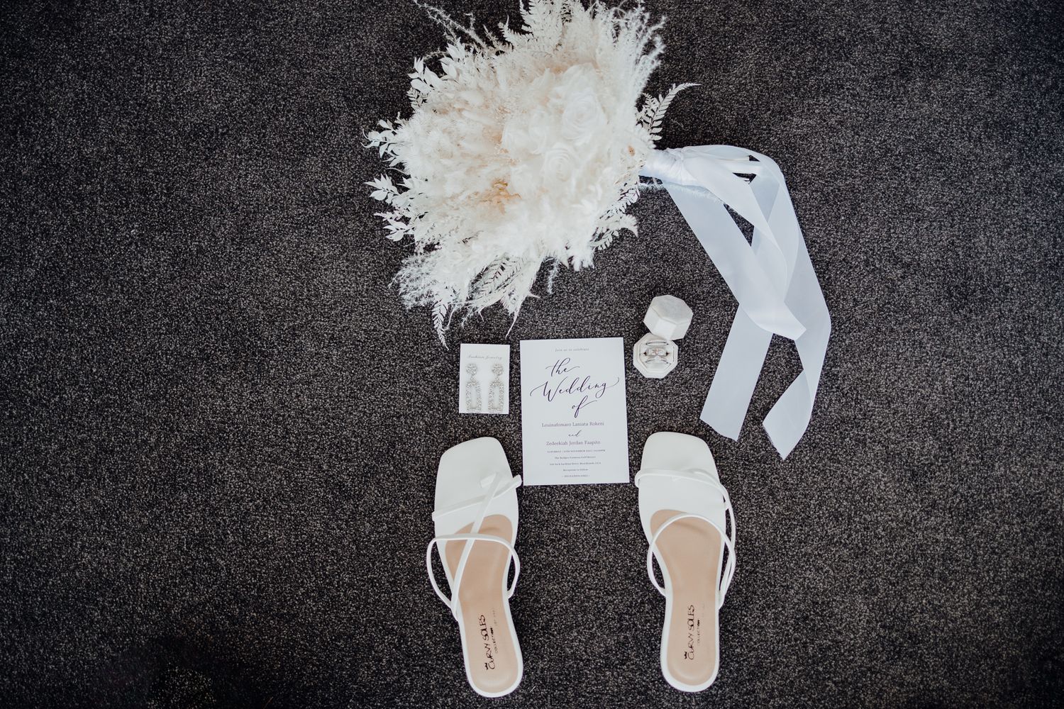 Wedding accessories including white sandals, dried flowers, and invitation laid out on dark fabric.