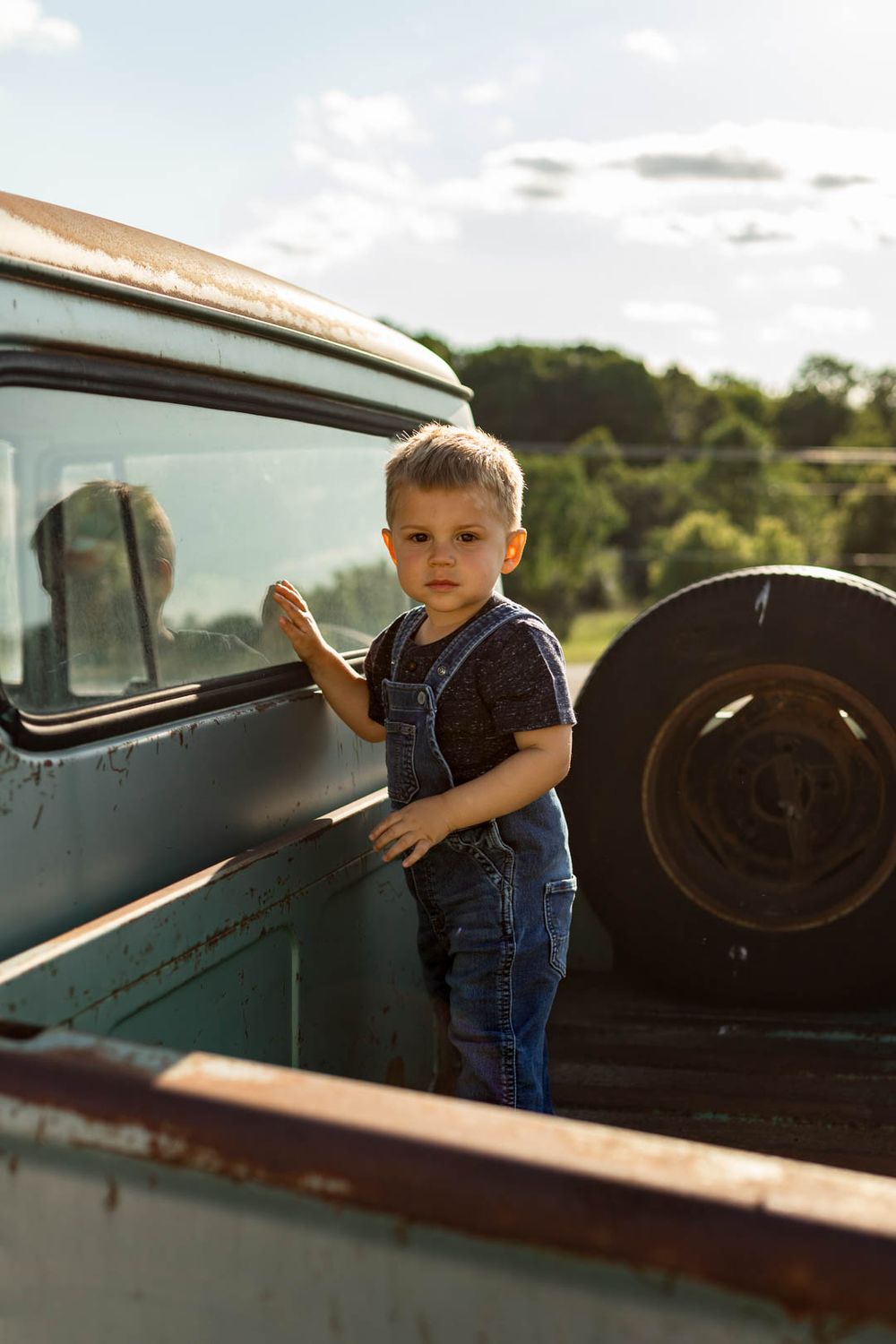 Small child in denim overalls standing in the bed of a vintage blue pickup truck.