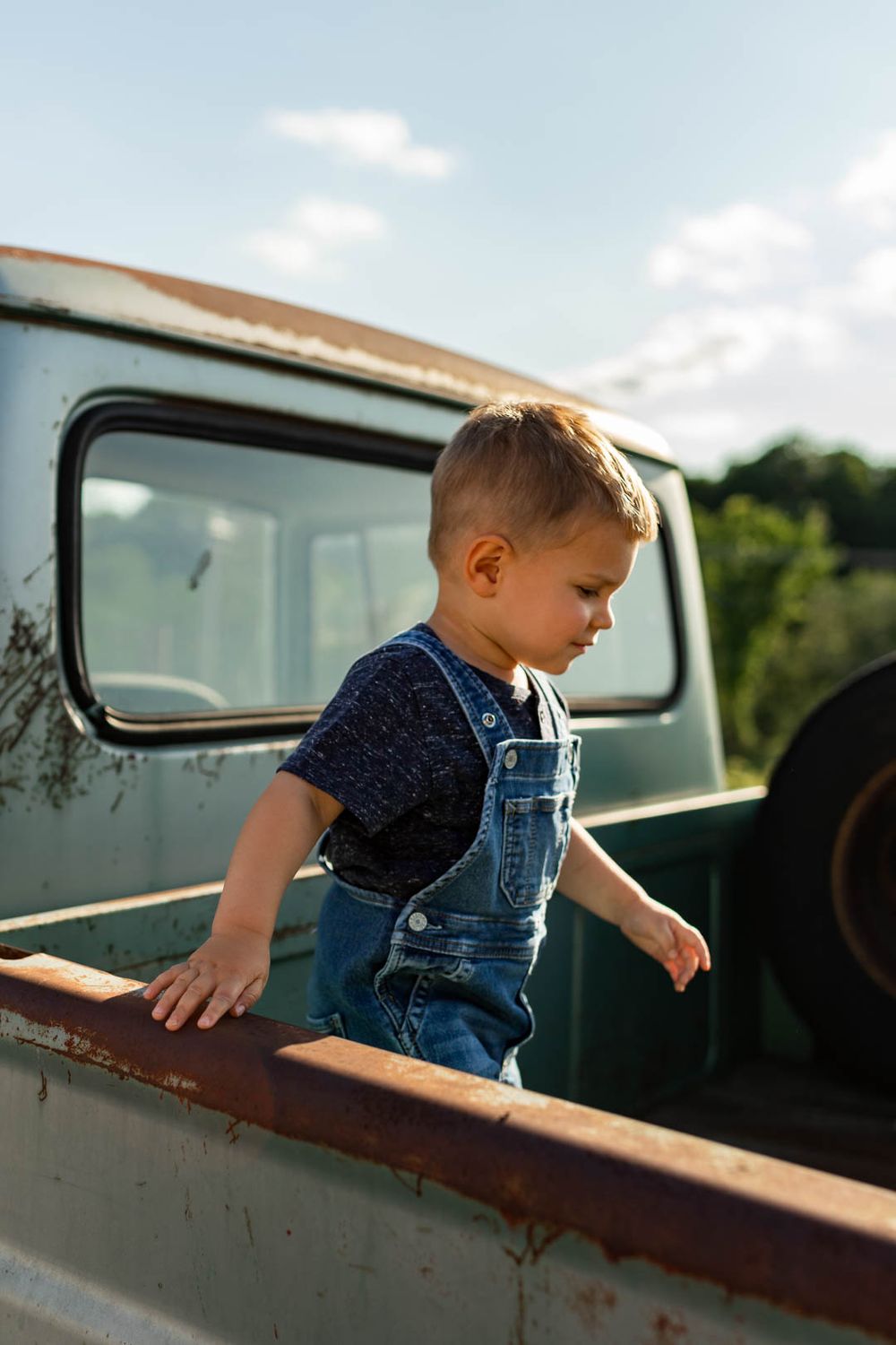 Young child in overalls exploring an old vintage truck bed on a sunny day.