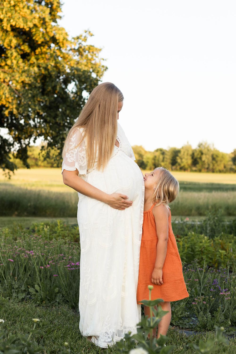 Maternity moment with woman in white dress and child in orange dress in summer field.