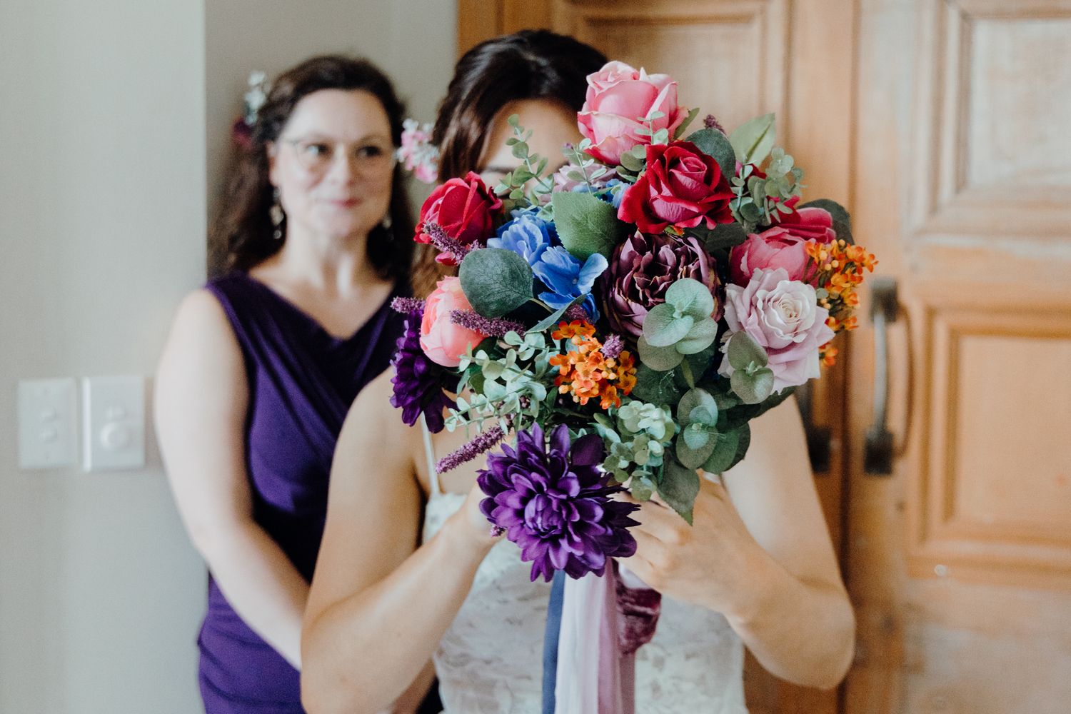 A vibrant multicolored wedding bouquet with roses, dahlias and greenery is held up against wooden doors.