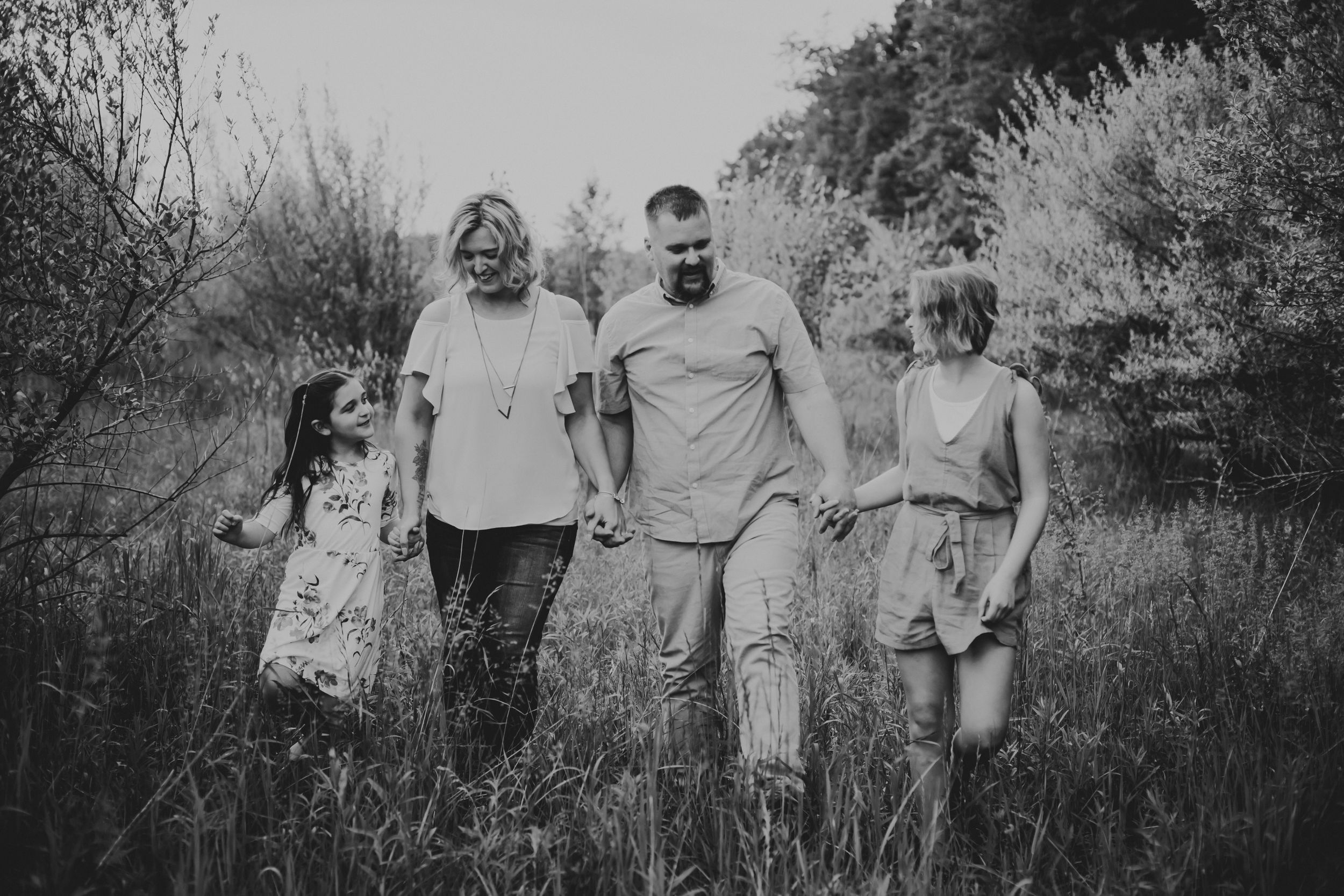 Black and white photo of mom, dad, and two daughters holding hands and walking through a field.