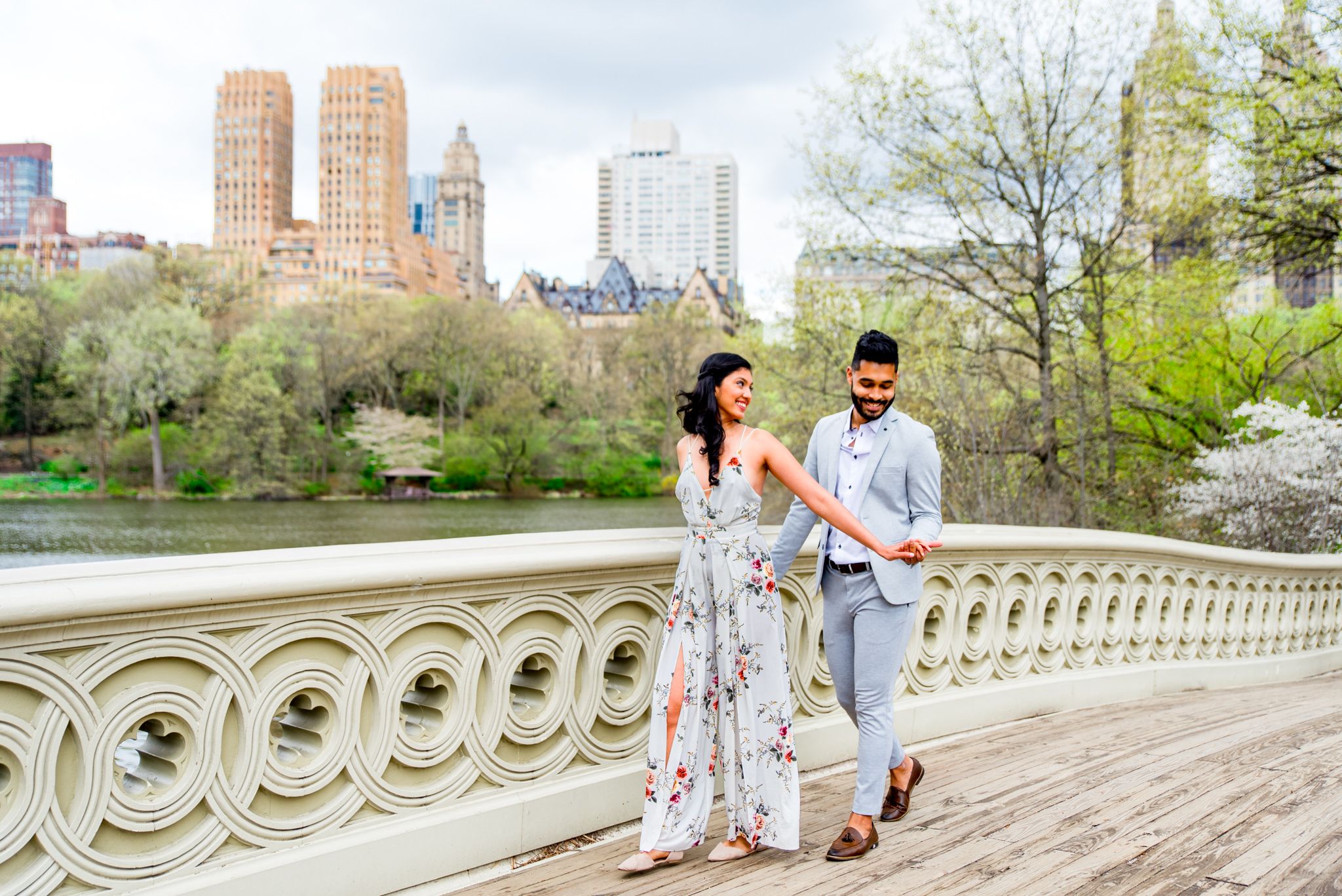 woman in floral jumpsuit leading man in grey suit across bow bridge for Central Park engagement photos