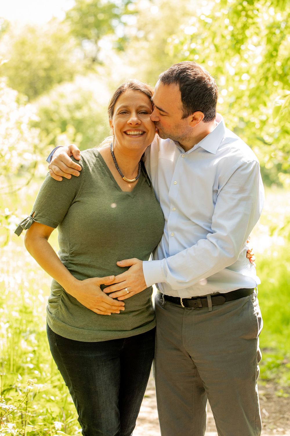 A couple share a hug in the park as the man holds his pregnant wife's stomach - Family Photography