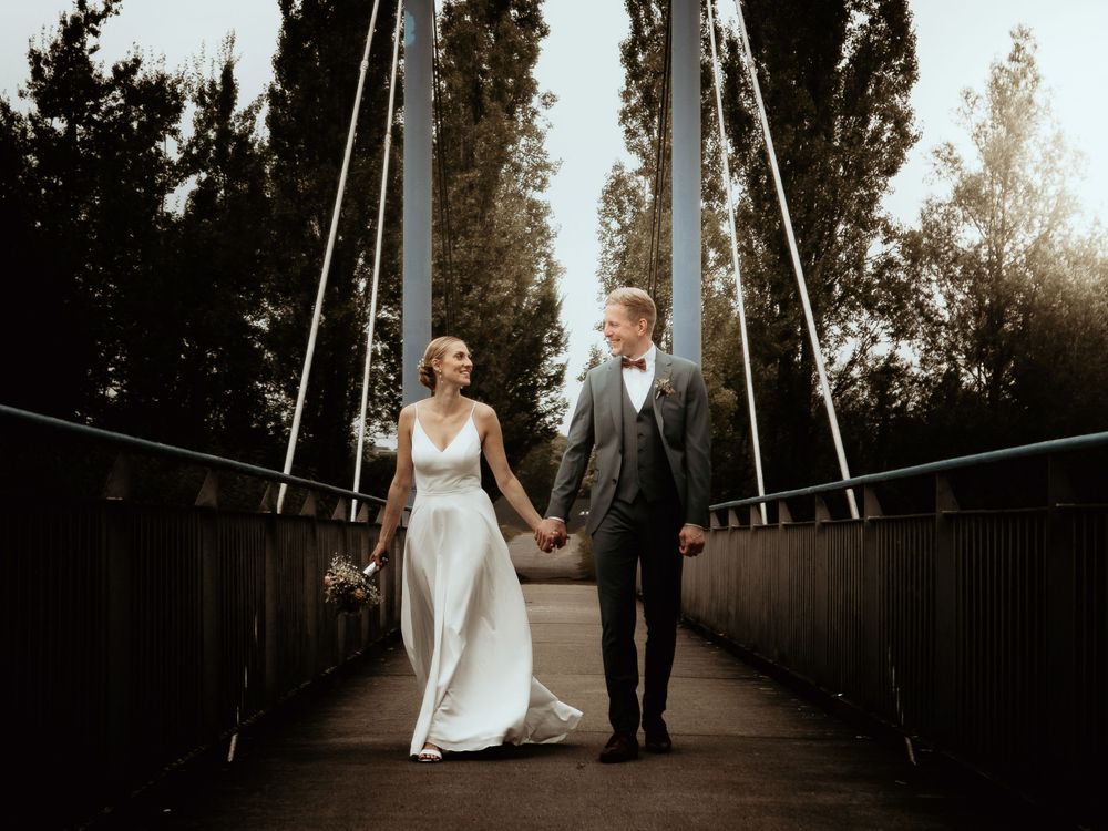 A couple in formal wedding attire walking hand in hand across a suspension bridge at sunset.