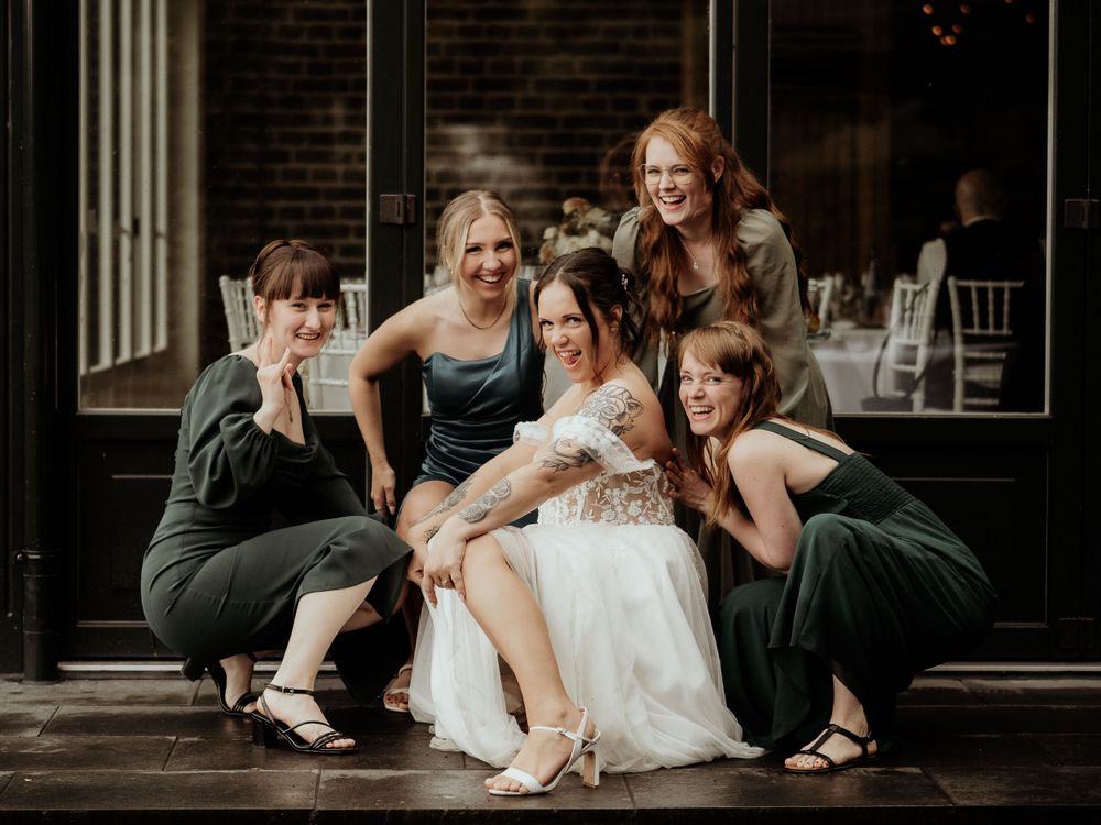 Women in green bridesmaid dresses crouch next to bride in white gown on wooden porch steps.