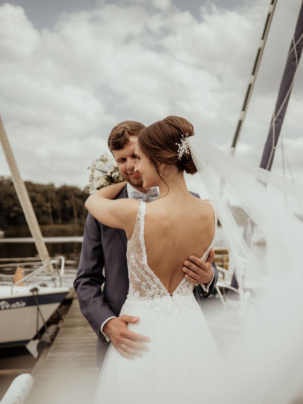 Newlywed couple embraces on dock with sailboats in background, bride wearing backless lace wedding dress.