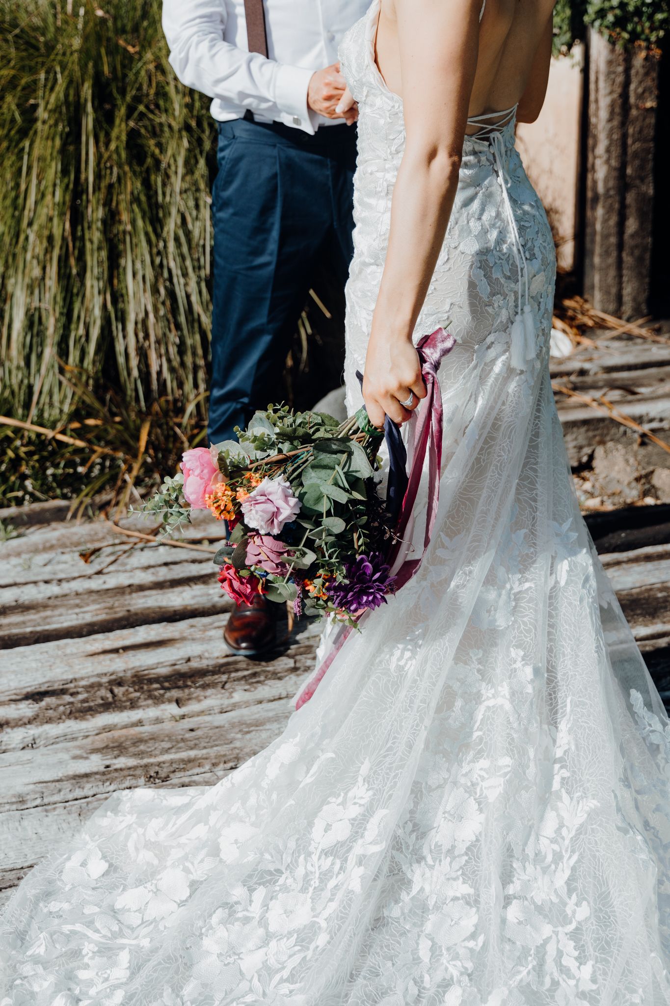 A close-up of a white lace wedding dress train and floral bouquet with pink and purple flowers.