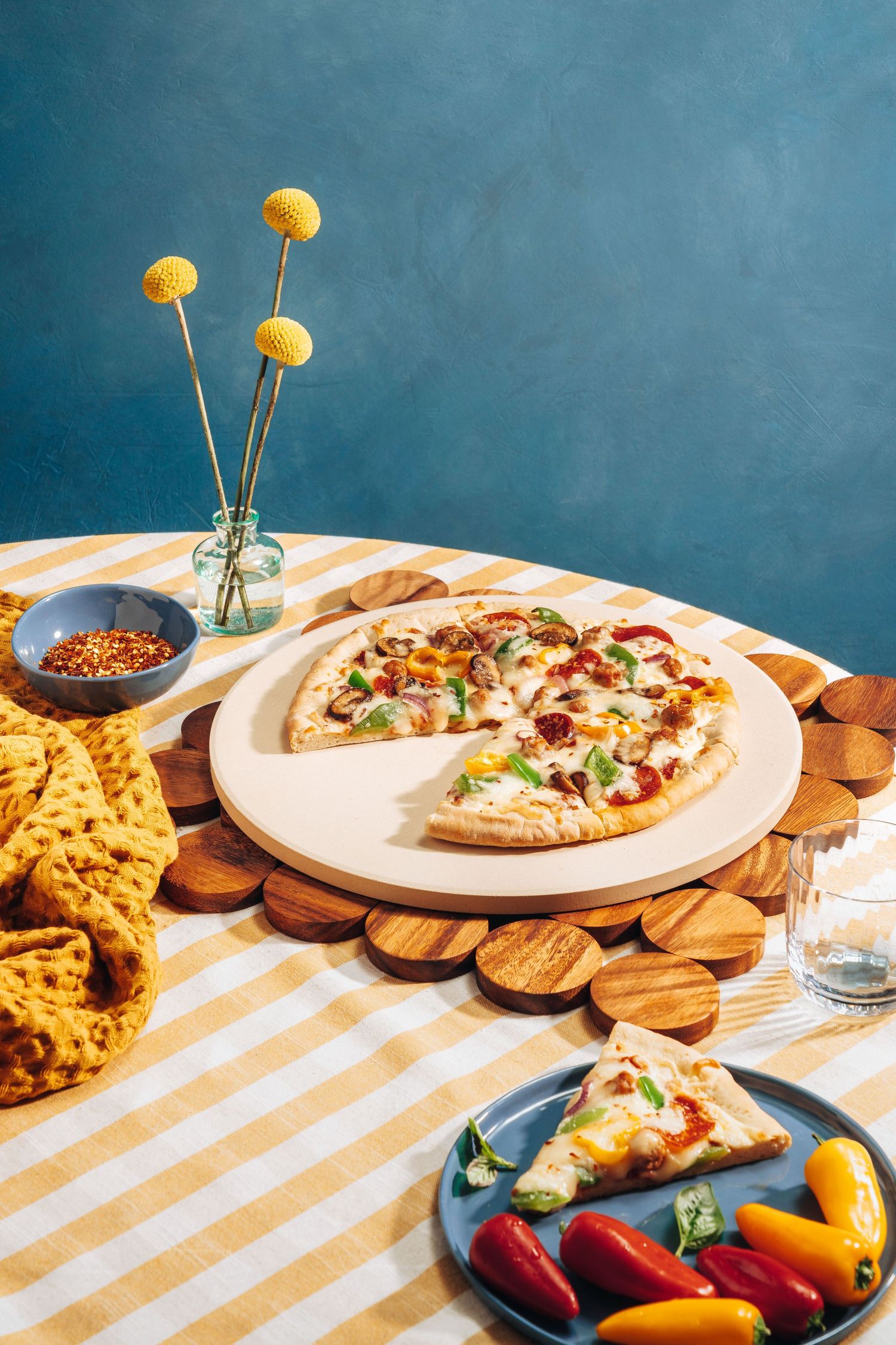 Pizza slice on white ceramic plate with yellow flowers and colorful peppers on striped tablecloth.